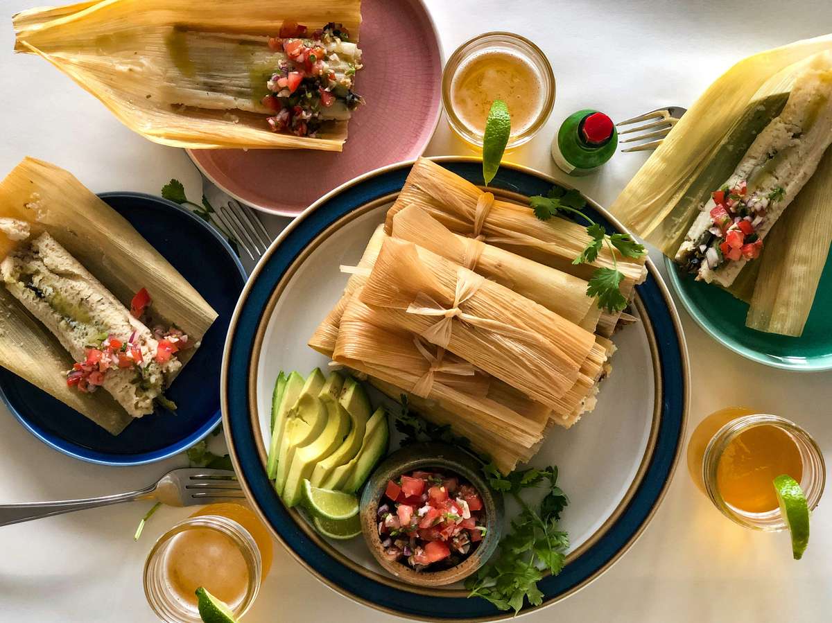 Traditional tamales wrapped in corn husks, served with avocado slices, pico de gallo, cilantro, and lime wedges on colorful plates.