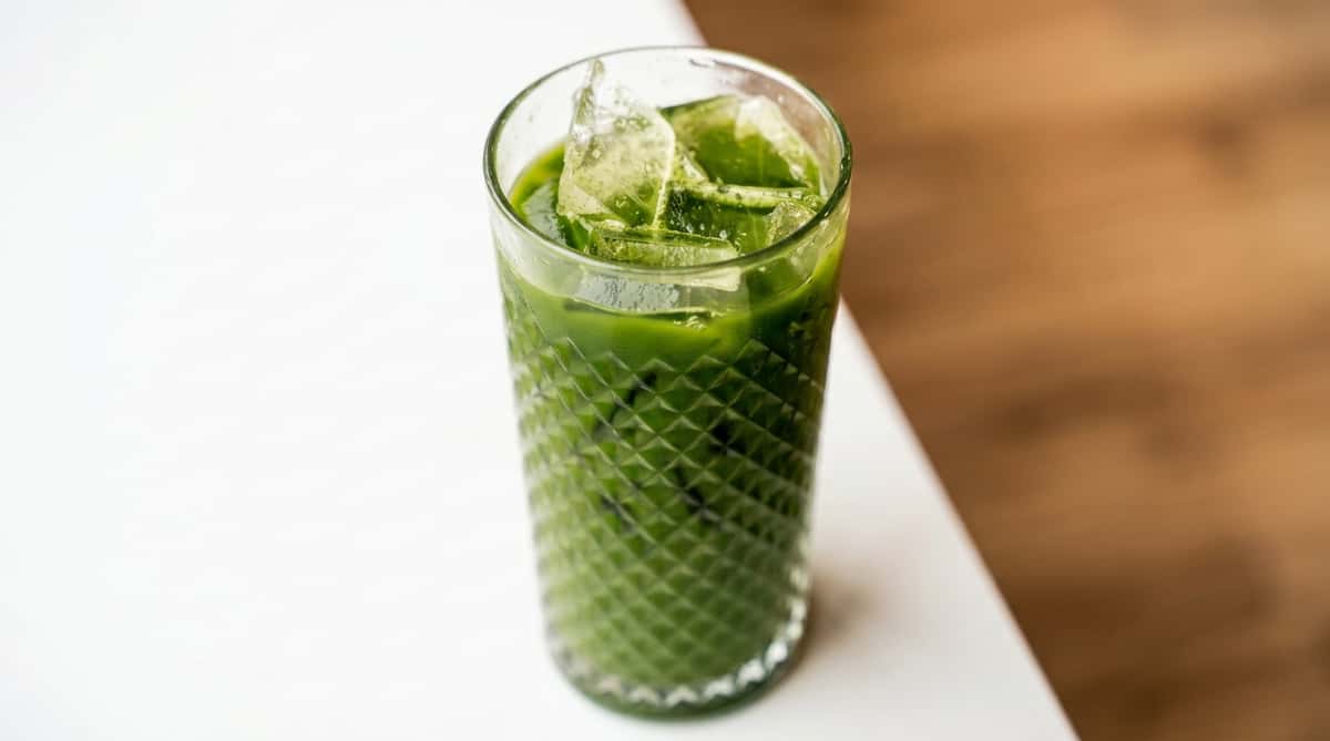 Iced matcha latte in a clear patterned glass with ice cubes, on a white surface.