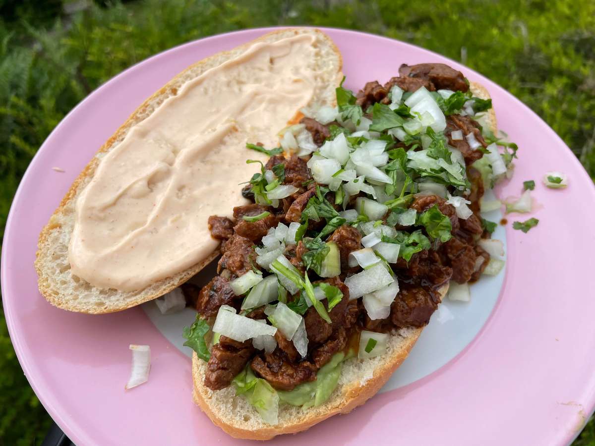 Open-faced sandwich on a pink plate, topped with chopped grilled meat, diced onions, fresh cilantro, guacamole, and a creamy sauce spread on the top half of the bread.