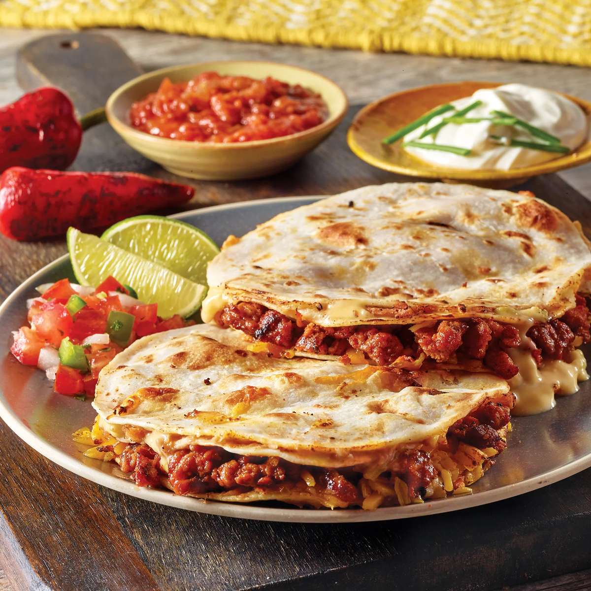 Plate with two cheese and chorizo quesadillas served with lime wedges, fresh pico de gallo, salsa, and a side of sour cream garnished with chives.