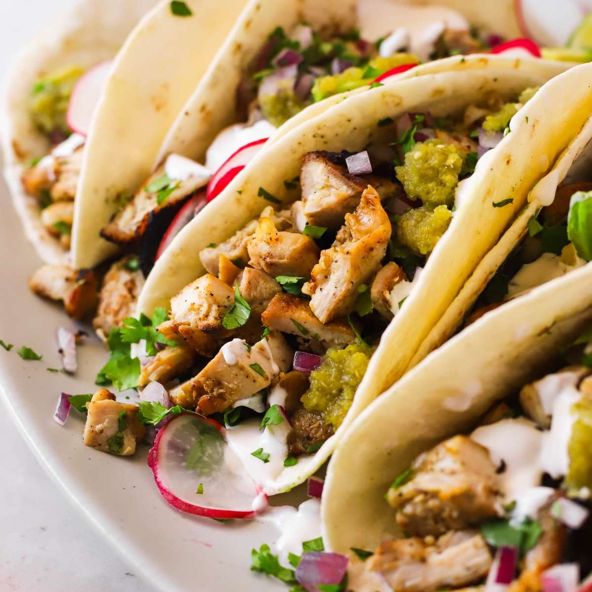 Close-up of soft chicken tacos topped with diced grilled chicken, green salsa, chopped red onions, fresh cilantro, sliced radishes, and a drizzle of crema on a white plate.