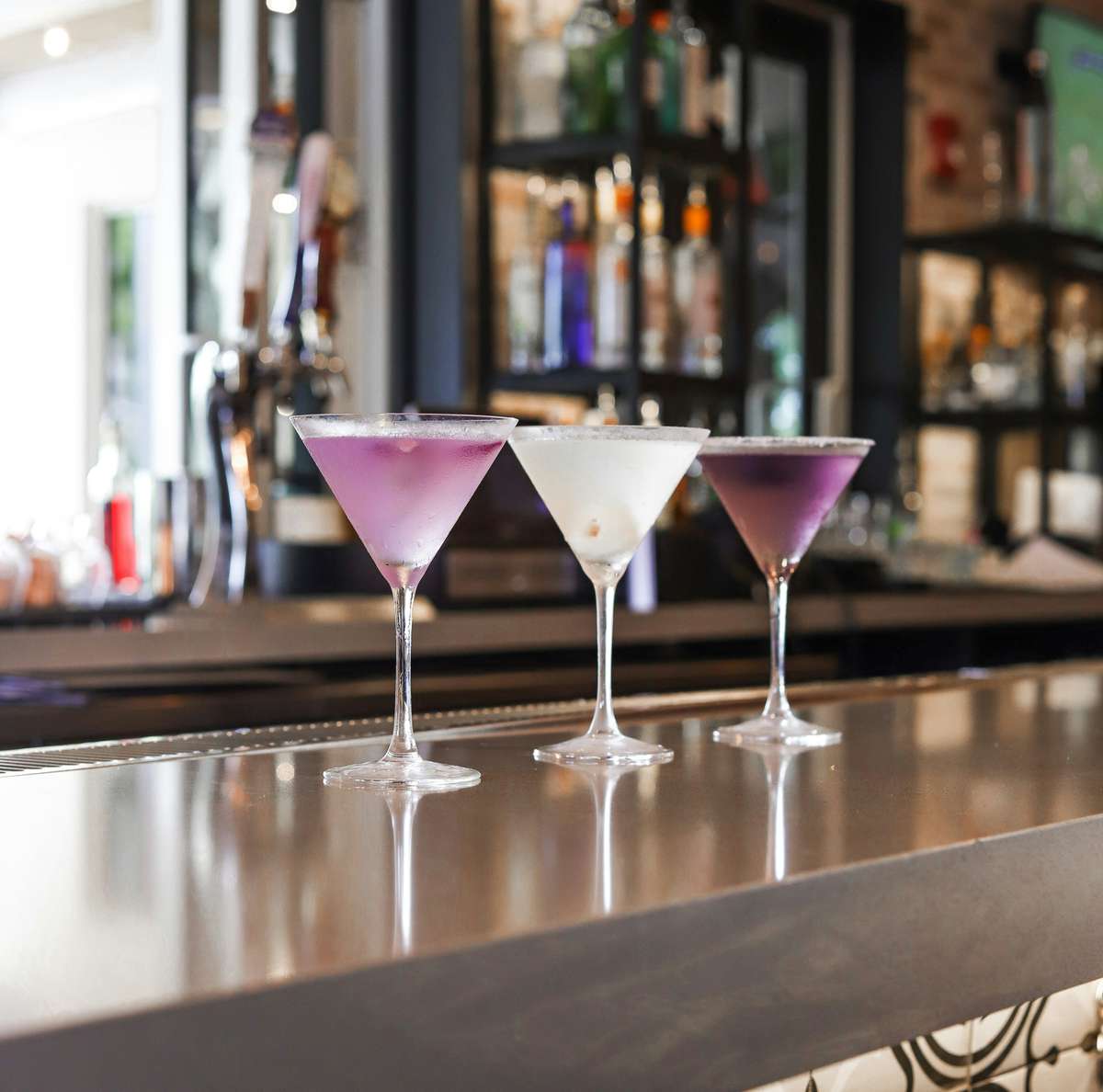 Three martini glasses with colorful cocktails—two purple and one white—elegantly displayed on a sleek bar counter.