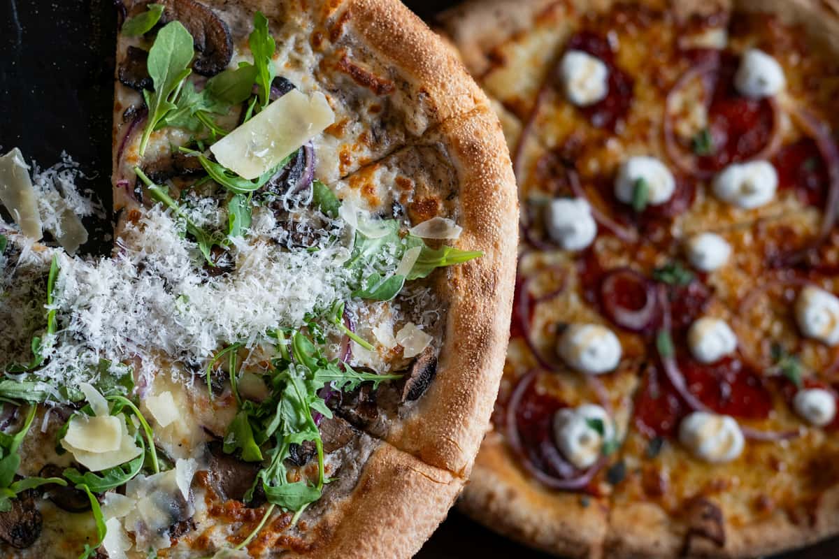 Mushroom arugula pizza topped with shaved parmesan next to a pepperoni pizza with ricotta and red onion.