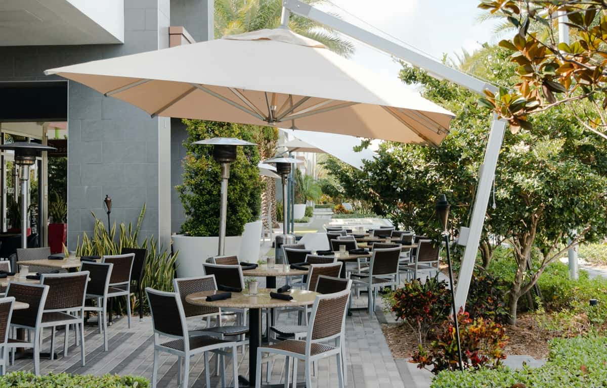Outdoor restaurant patio with wicker chairs, round tables, large umbrellas, and lush greenery creating a relaxed dining atmosphere.
