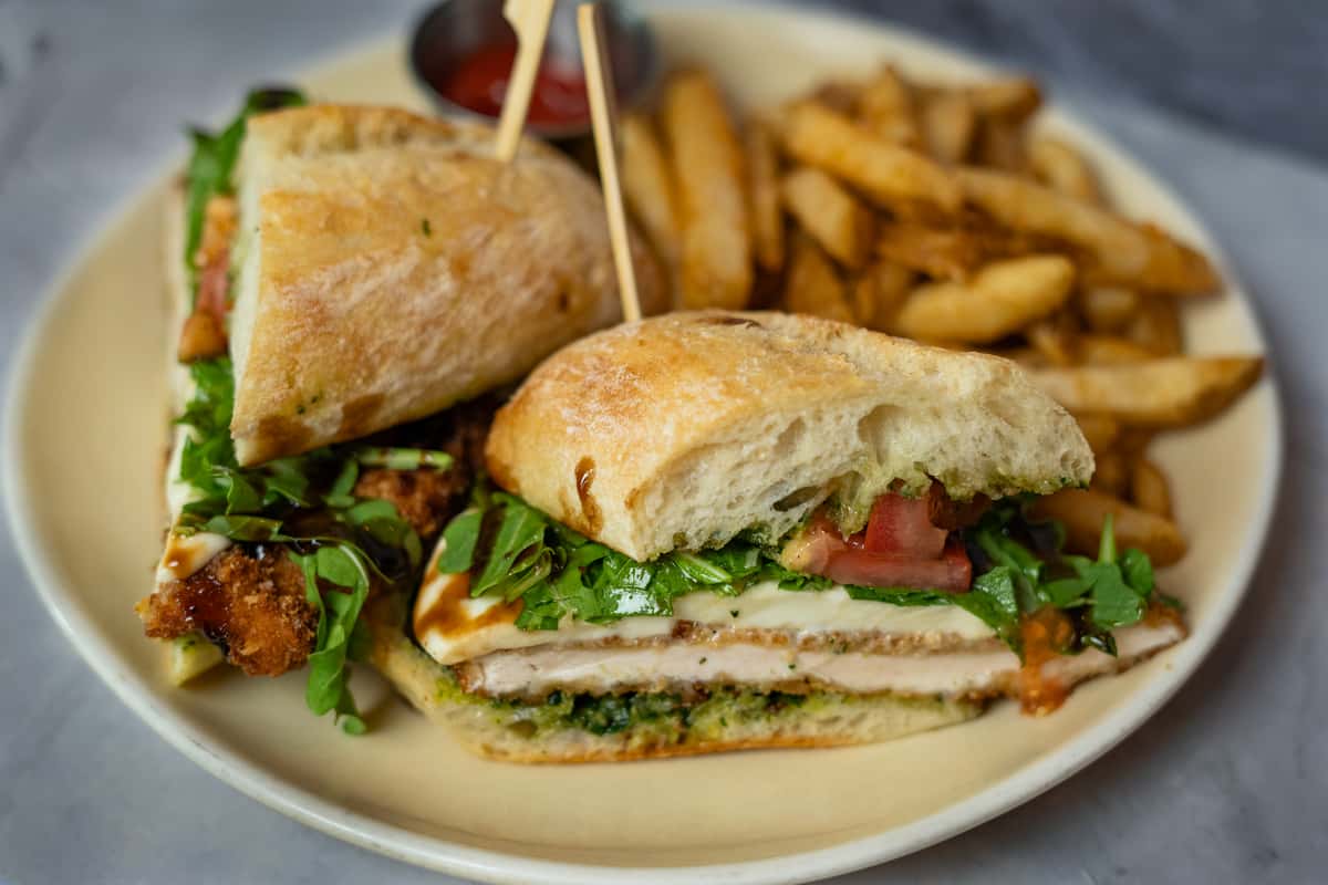 Crispy chicken sandwich with lettuce, tomato, and pesto on ciabatta bread served with a side of golden fries and ketchup.