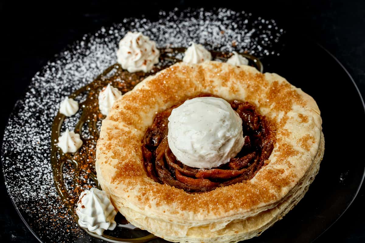 Caramelized apple tart with a puff pastry crust, topped with vanilla ice cream, dollops of whipped cream, caramel drizzle, and powdered sugar.