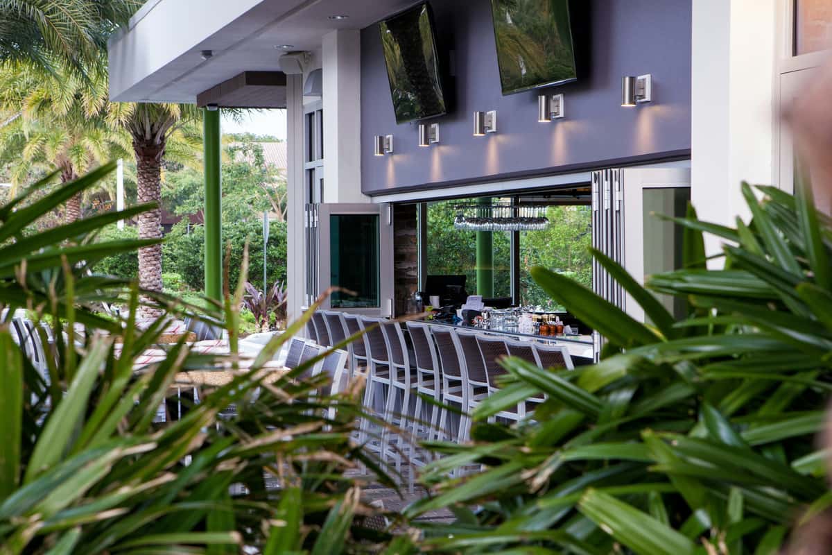 Modern outdoor bar with high chairs, flat-screen TVs, and tropical greenery surrounding the seating area.