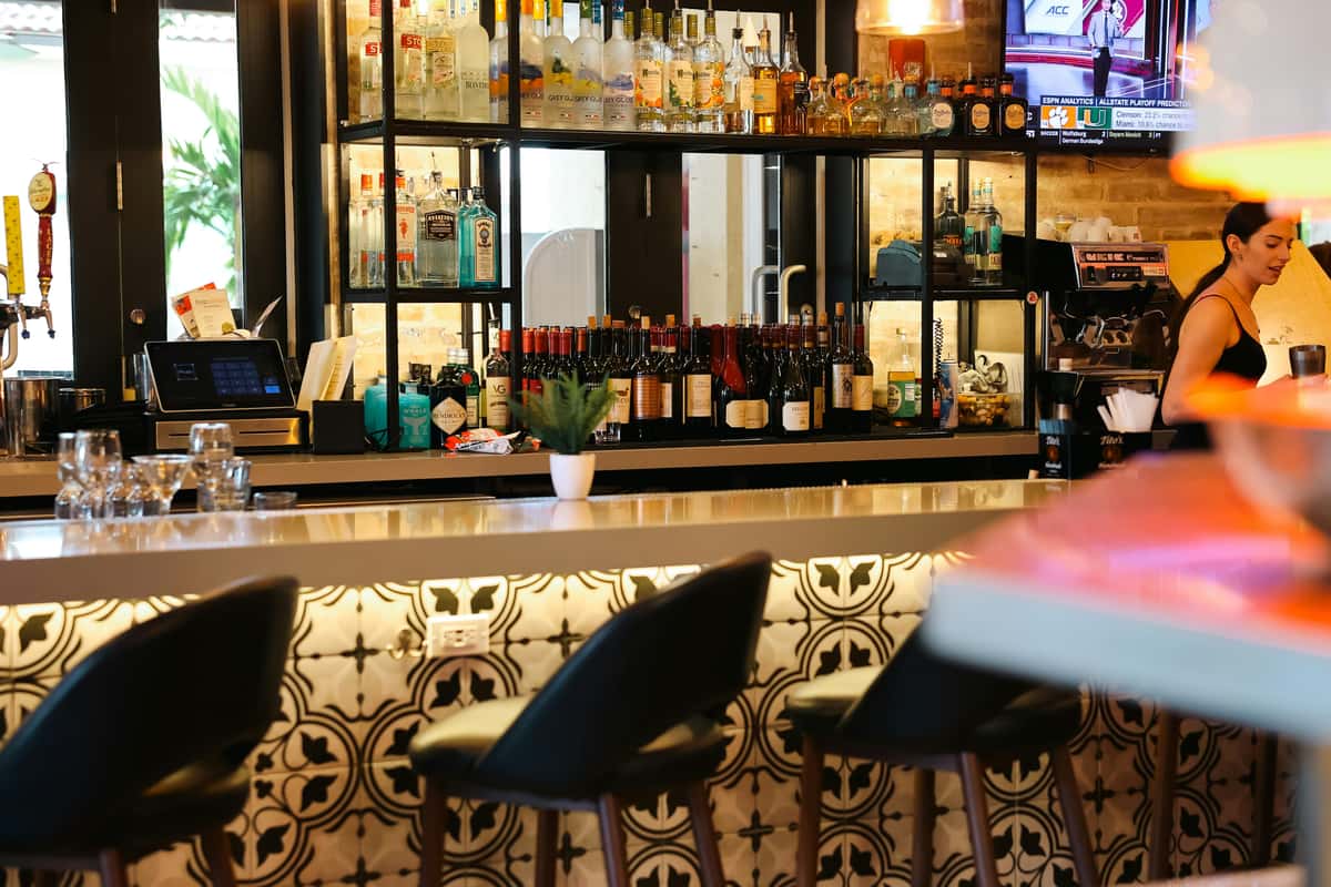 Stylish indoor bar with patterned tile, stocked shelves of liquor and wine, and a bartender preparing a drink.