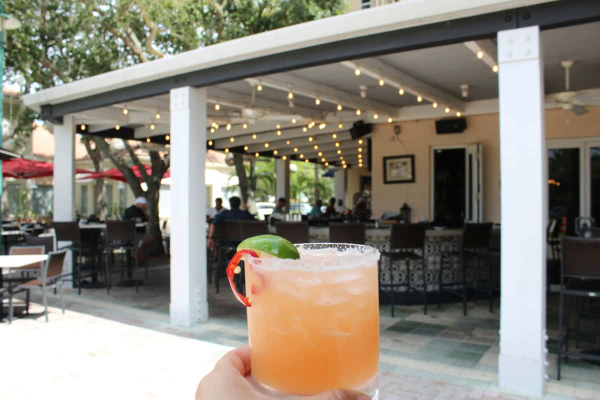Iced margarita garnished with lime and chili held in front of a covered outdoor patio with string lights and bar seating.