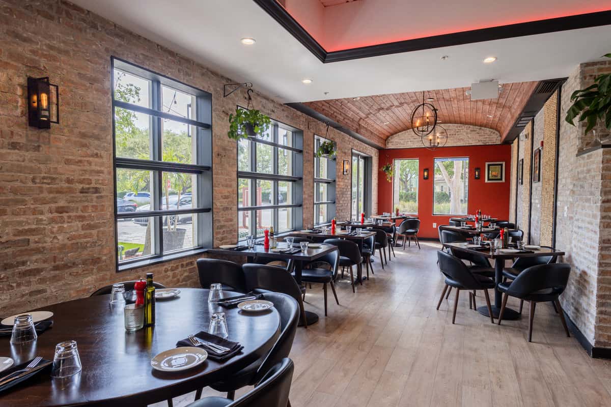 Cozy dining room with brick walls, large windows, black chairs, wood tables, and warm lighting under a vaulted wooden ceiling.