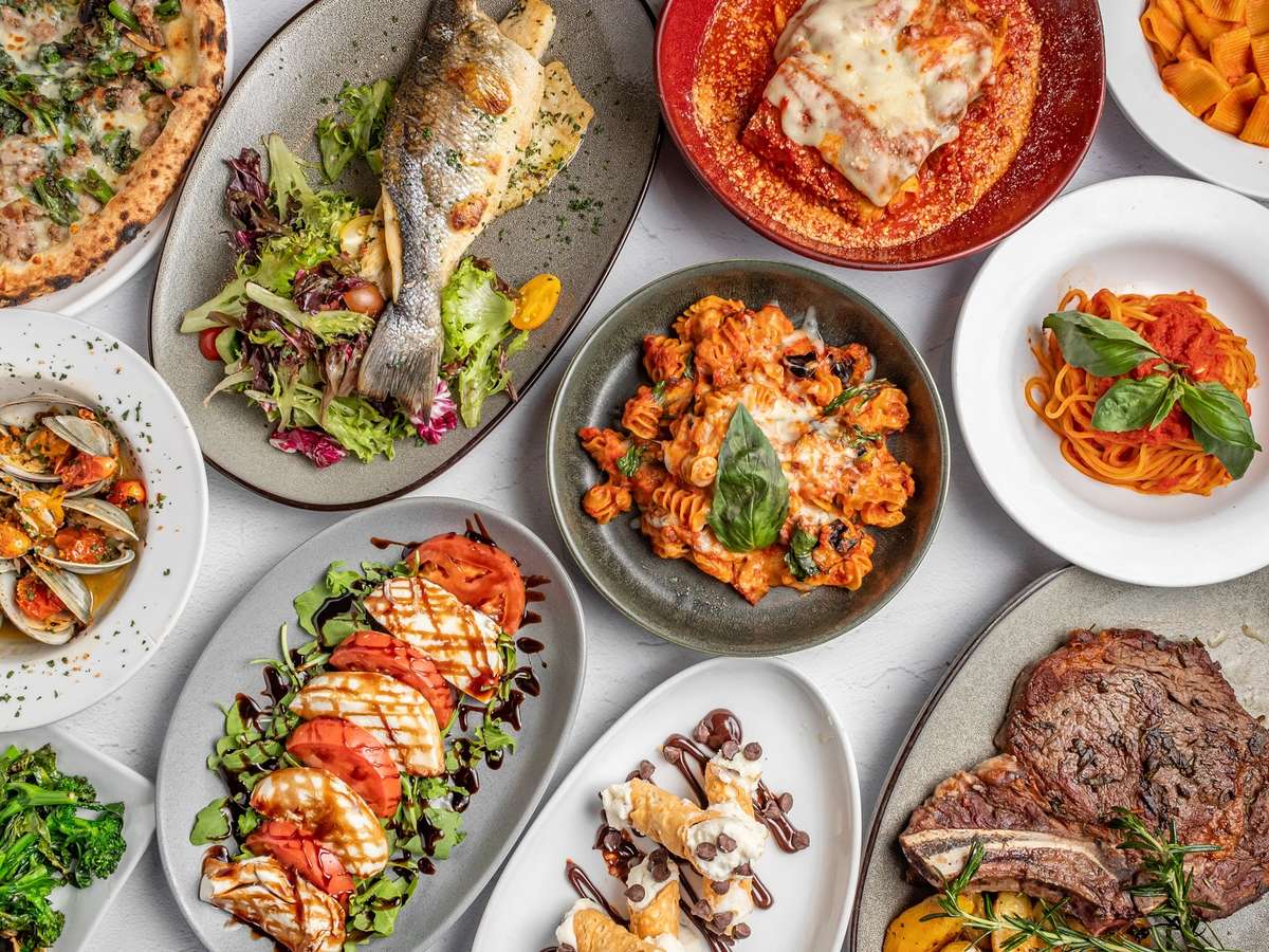 Italian feast with lasagna, seafood, grilled fish, steak, caprese salad, and various pasta dishes arranged on a white table.