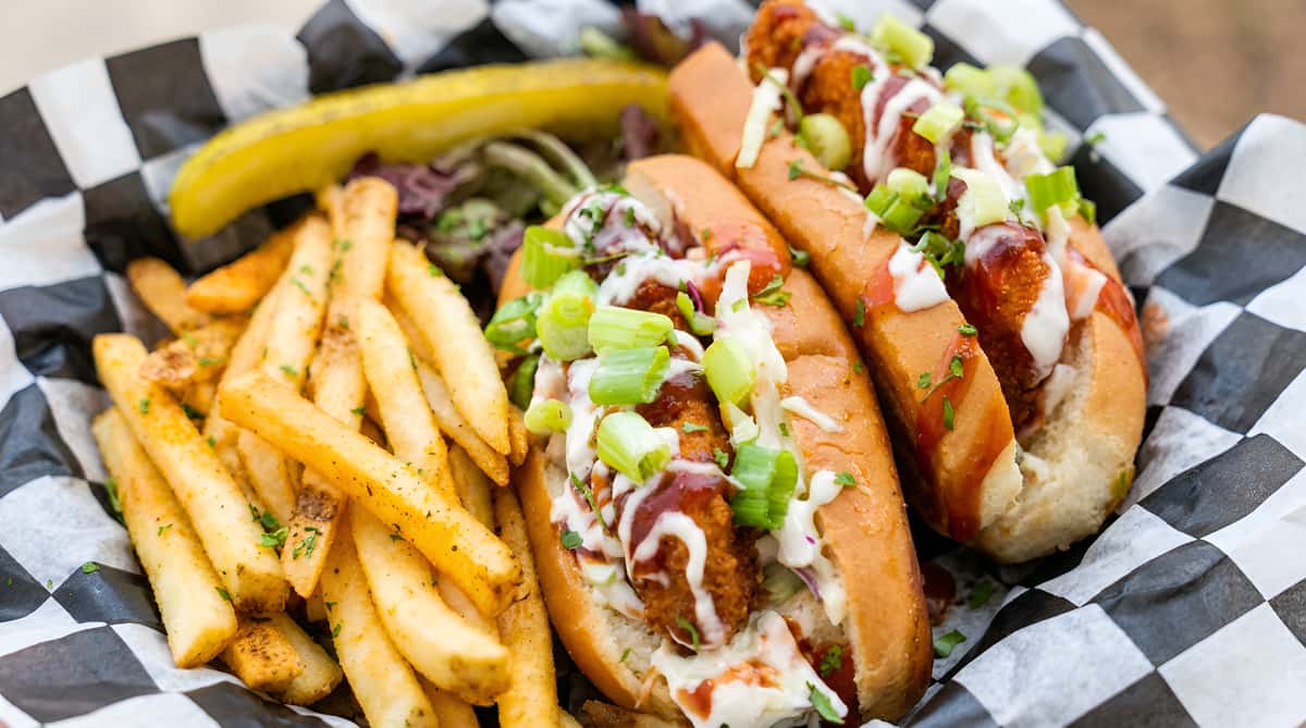 Two hot dog buns filled with fried food, coleslaw, sauces, and scallions, served with fries and a pickle.