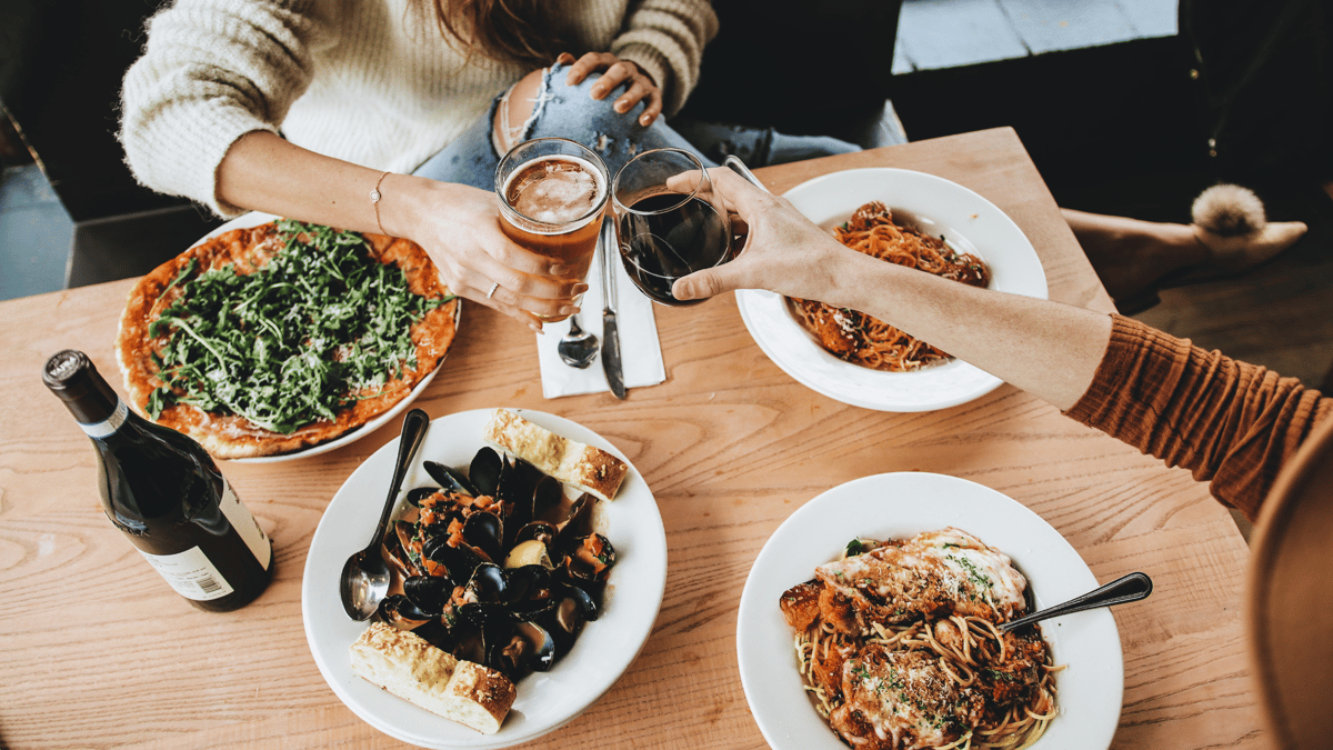 Neighbors clinking beer and wine glasses over a delicious spread of Osteria's Italian pasta and pizza classics