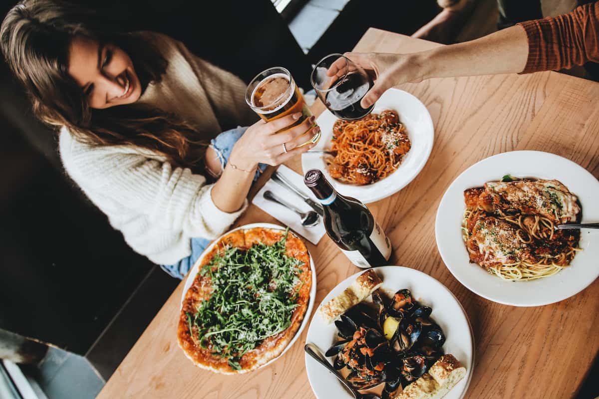 Two people toasting drinks over a table full of Italian dishes including pasta, mussels, and arugula pizza.