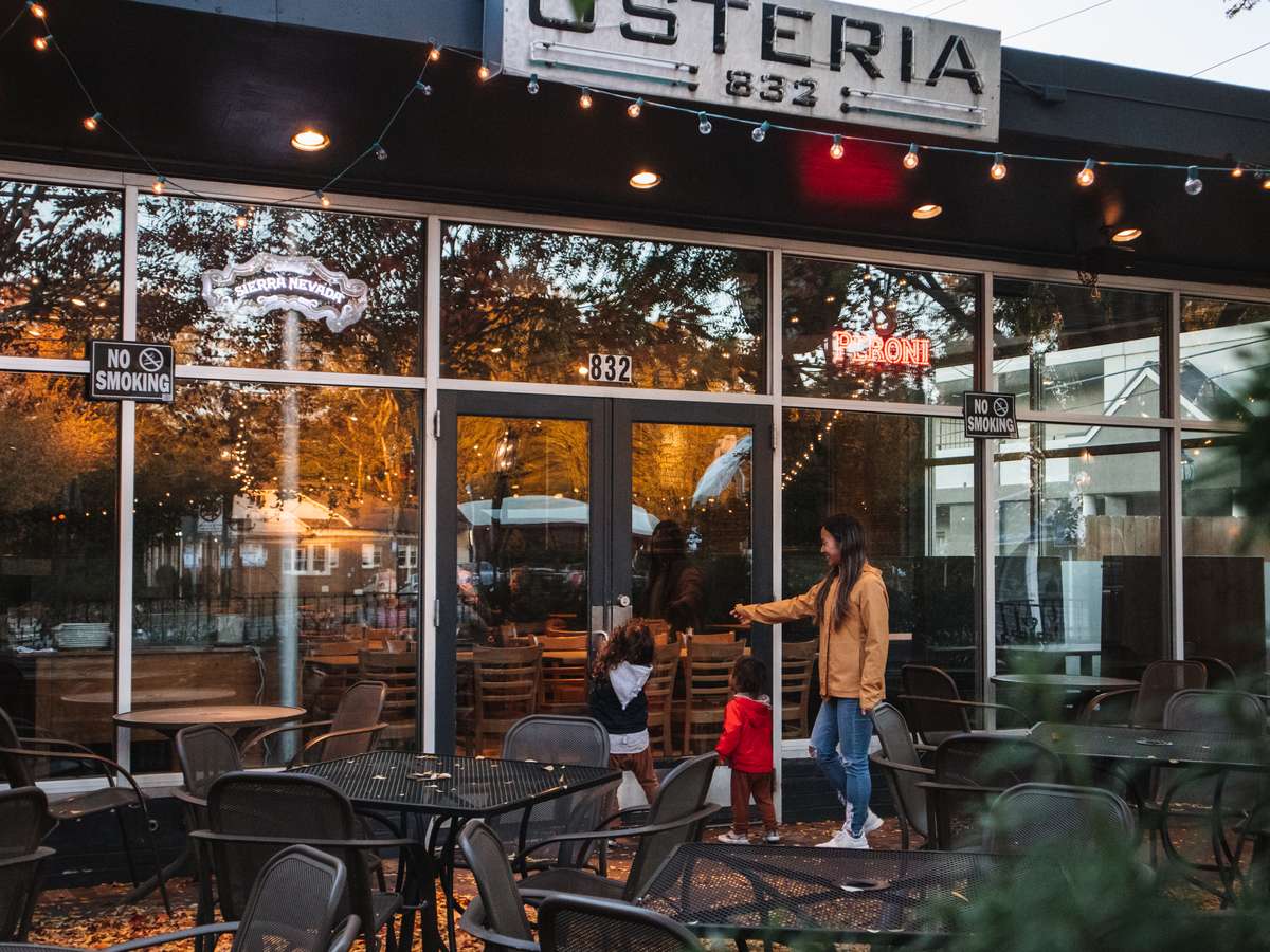 Exterior of Osteria 832 restaurant with string lights, outdoor seating, and a family entering through the glass doors.