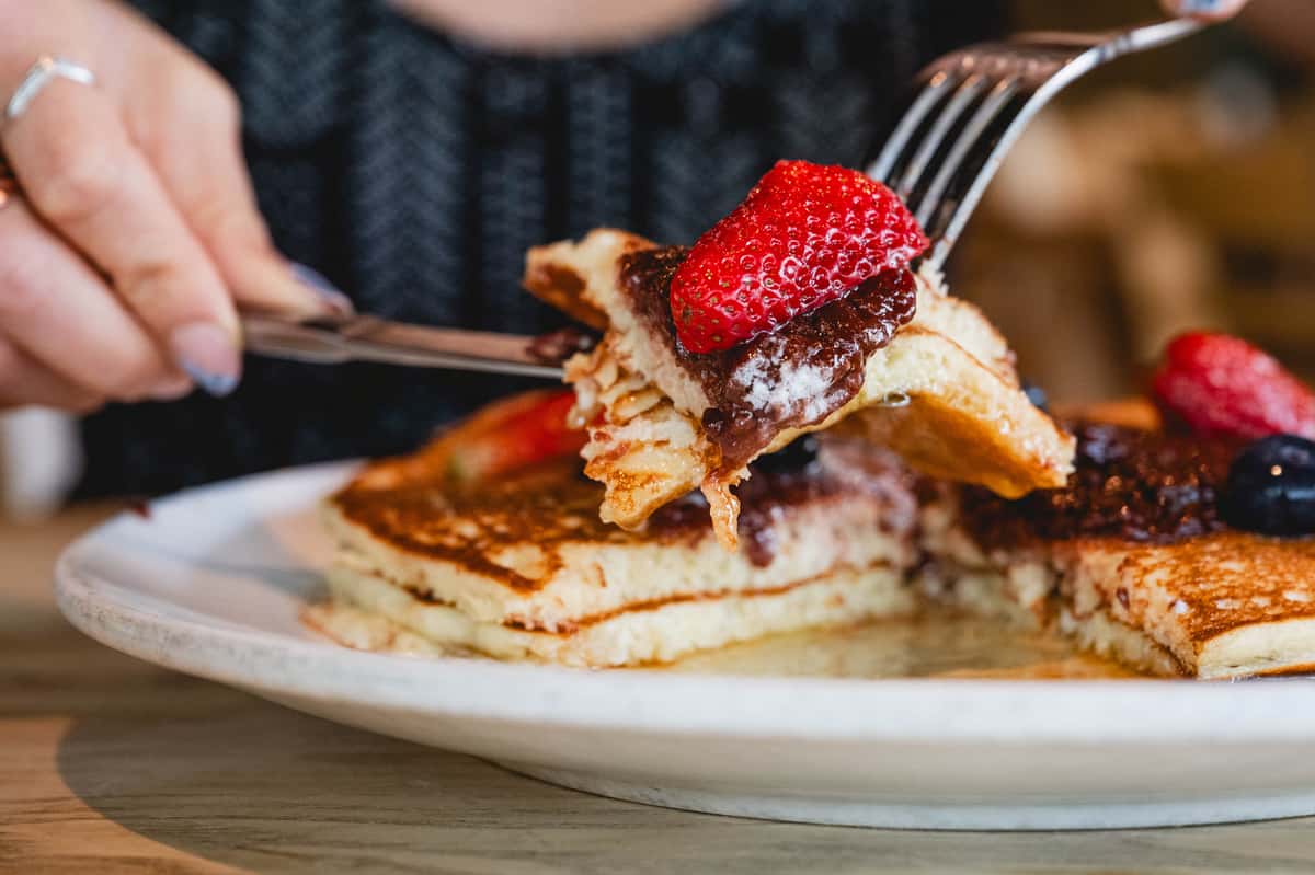 Fluffy pancakes topped with chocolate spread, fresh strawberries, and blueberries being cut and served on a white plate.