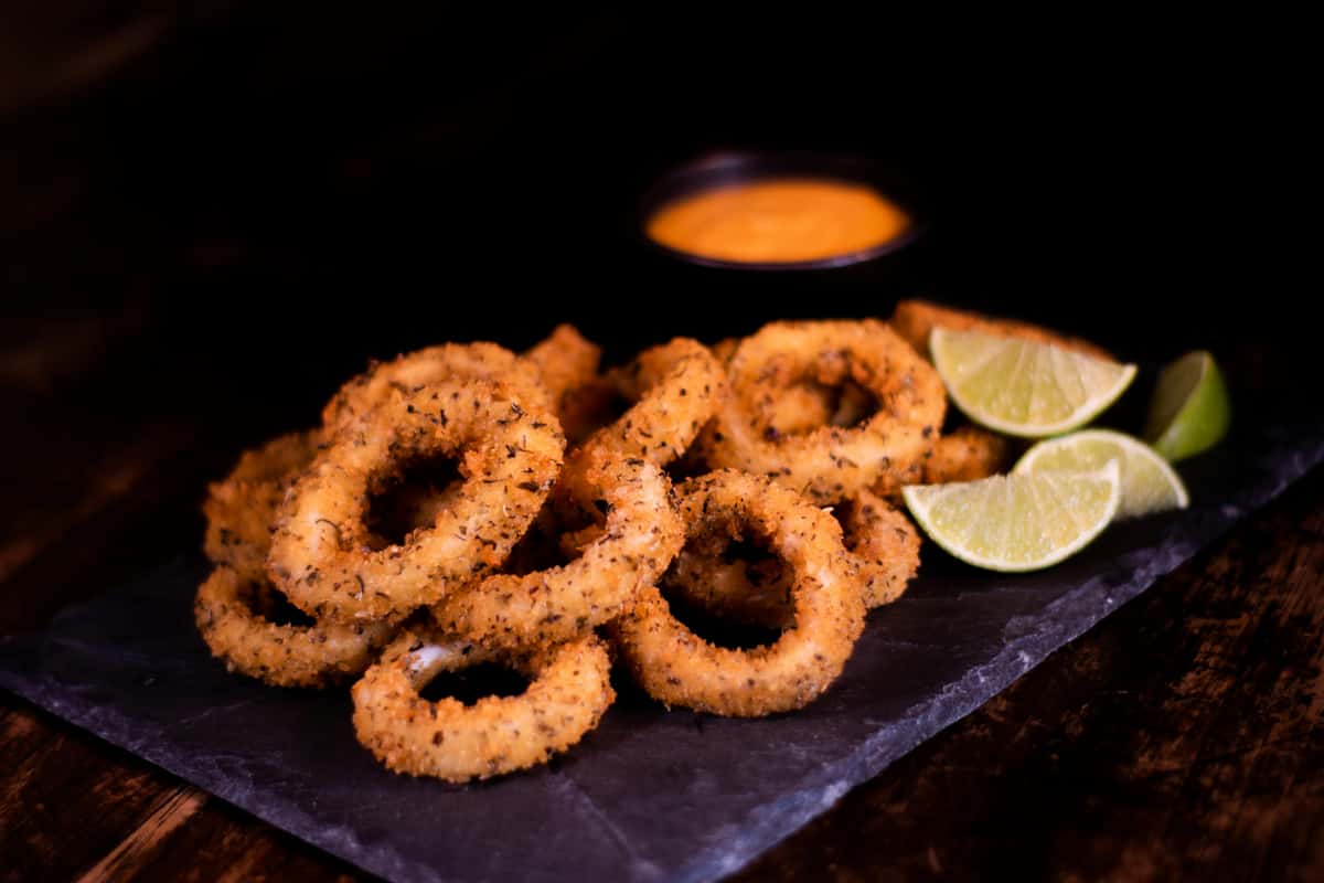 Crispy herb-seasoned fried calamari rings served on a slate board with lime wedges and dipping sauce—golden seafood appetizer with a rustic restaurant presentation.