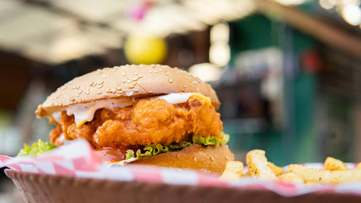 Crispy fried chicken sandwich on a sesame seed bun with lettuce and creamy sauce, served with seasoned fries—golden, crunchy comfort food in a casual street-style setting.