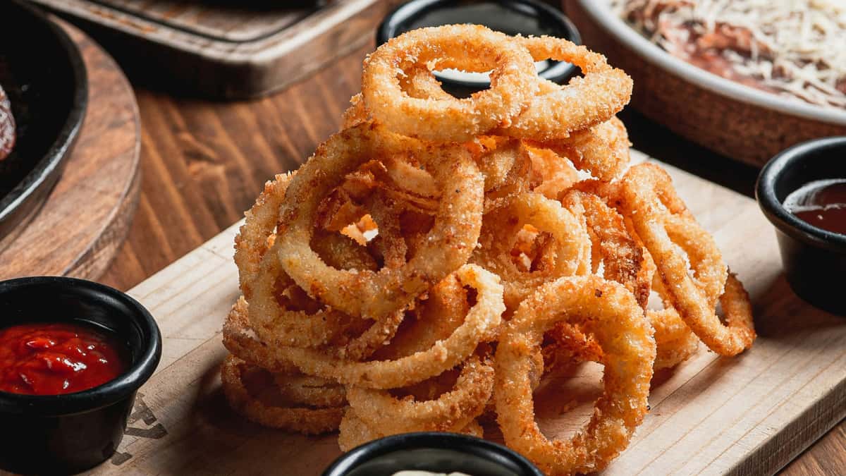 Crispy golden onion rings stacked on a wooden board, served with dipping sauces—classic bar snack with crunchy breaded coating and rustic presentation.