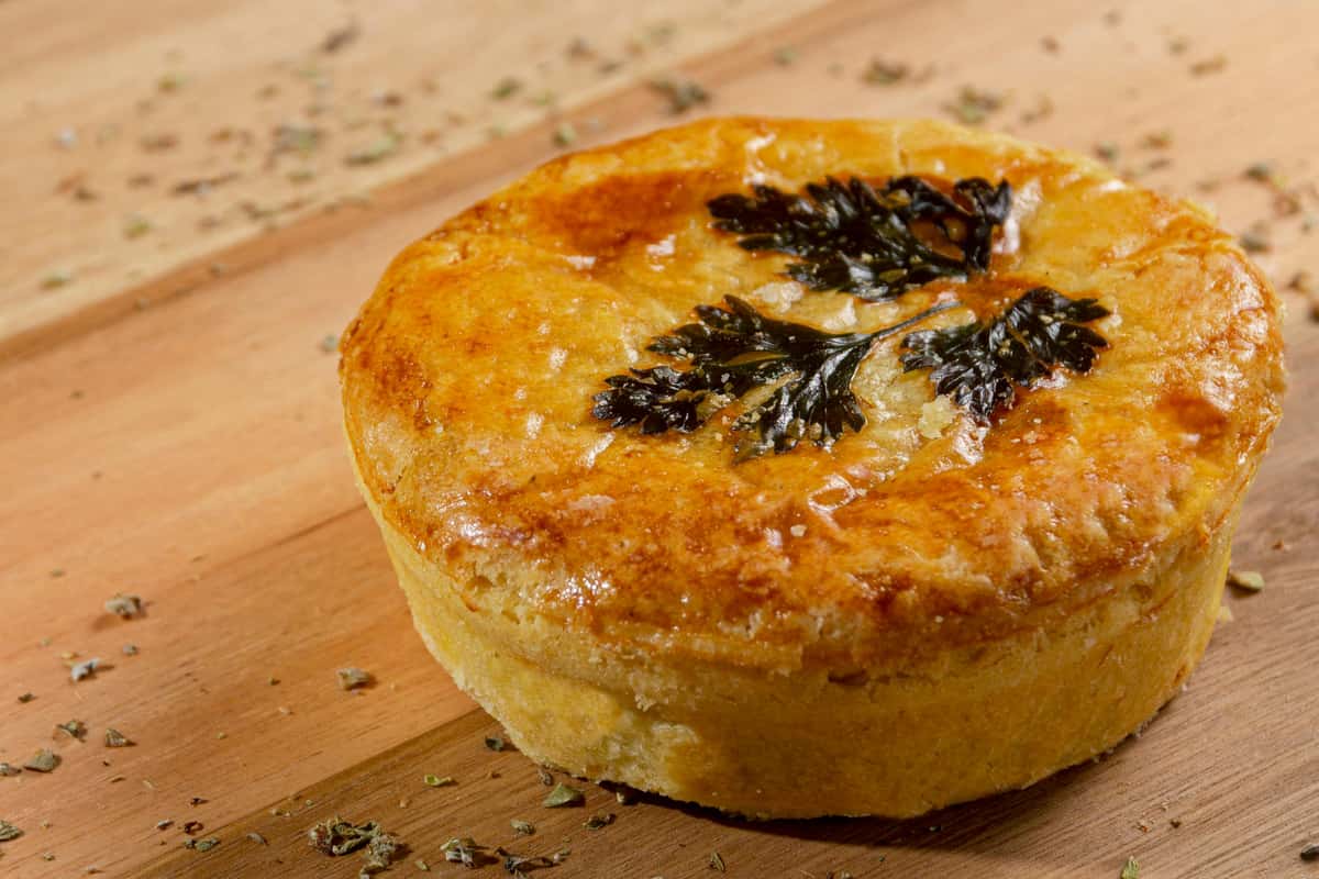 Golden-brown individual savory pie with a flaky crust, topped with herbs and baked to a glossy finish—rustic comfort food served on a wooden board.