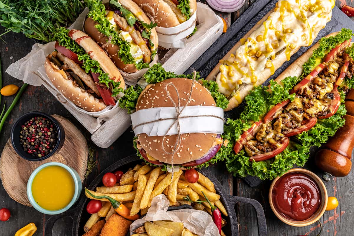 A variety of delicious fast foods on a wooden platter, featuring a large sesame seed hamburger, hot dogs, french fries, and fresh greens.