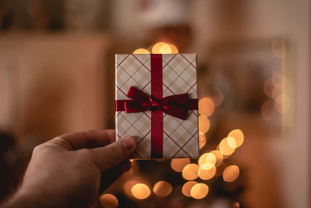 Hand holding a small, neatly wrapped gift with a red ribbon in front of warm, festive bokeh lights.