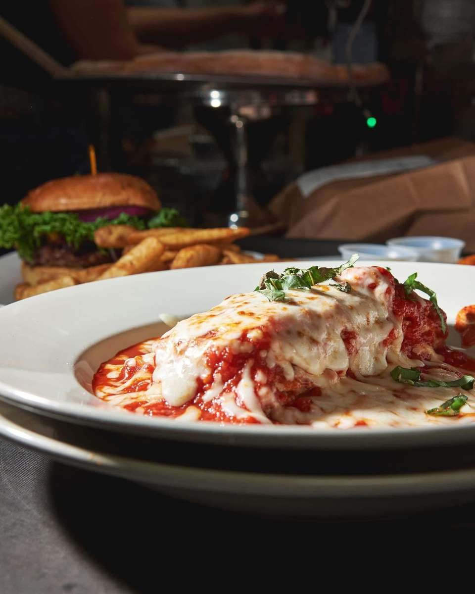 Close-up of a rich, oven-baked lasagna smothered in melted mozzarella cheese and savory marinara sauce, garnished with fresh basil, served at a restaurant with a classic cheeseburger and fries visible in the background.