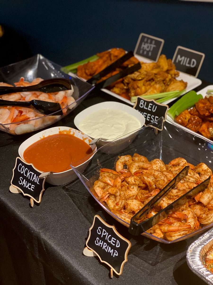 Buffet setup with chilled shrimp and spicy seasoned shrimp alongside cocktail sauce, blue cheese dip, and platters of mild and hot buffalo wings with celery sticks.