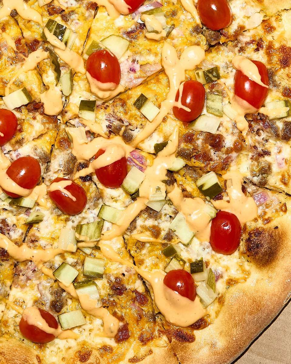 Close-up of a cheeseburger-inspired pizza topped with melted cheddar, ground beef, diced pickles, red onions, cherry tomatoes, and a drizzle of creamy burger sauce on a golden-brown crust.