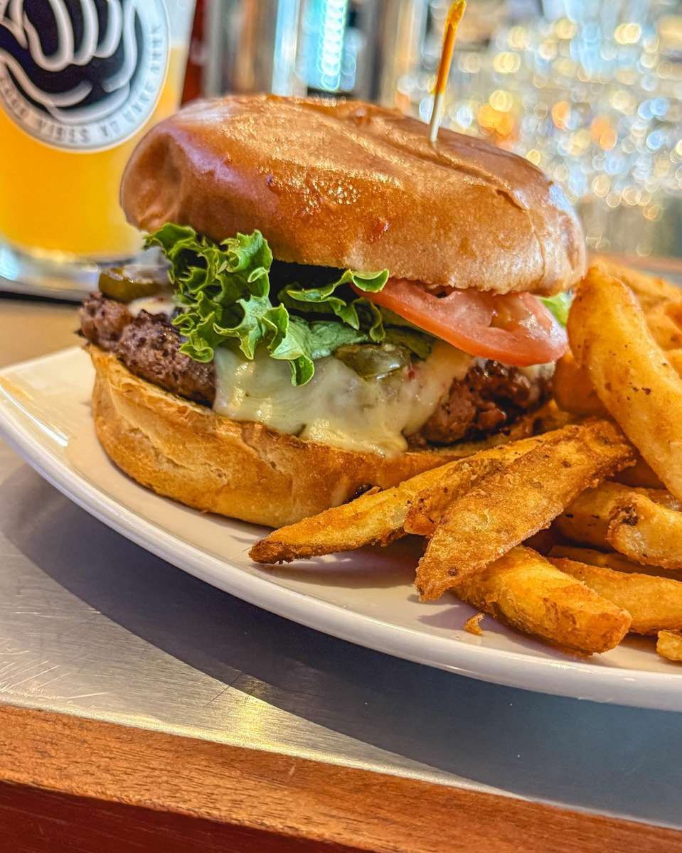Juicy cheeseburger topped with melted Swiss cheese, lettuce, tomato, and pickles on a toasted brioche bun, served with seasoned fries and a cold craft beer in a casual bar setting.