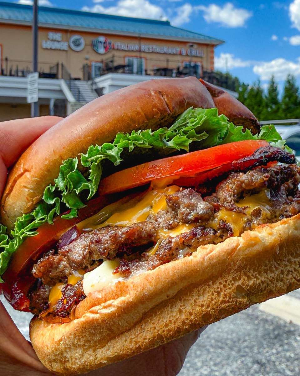 Close-up of a juicy double cheeseburger stacked with melted cheddar cheese, fresh lettuce, sliced tomato, onions, and grilled beef patties on a toasted brioche bun, held in front of a casual American restaurant under a bright blue sky.