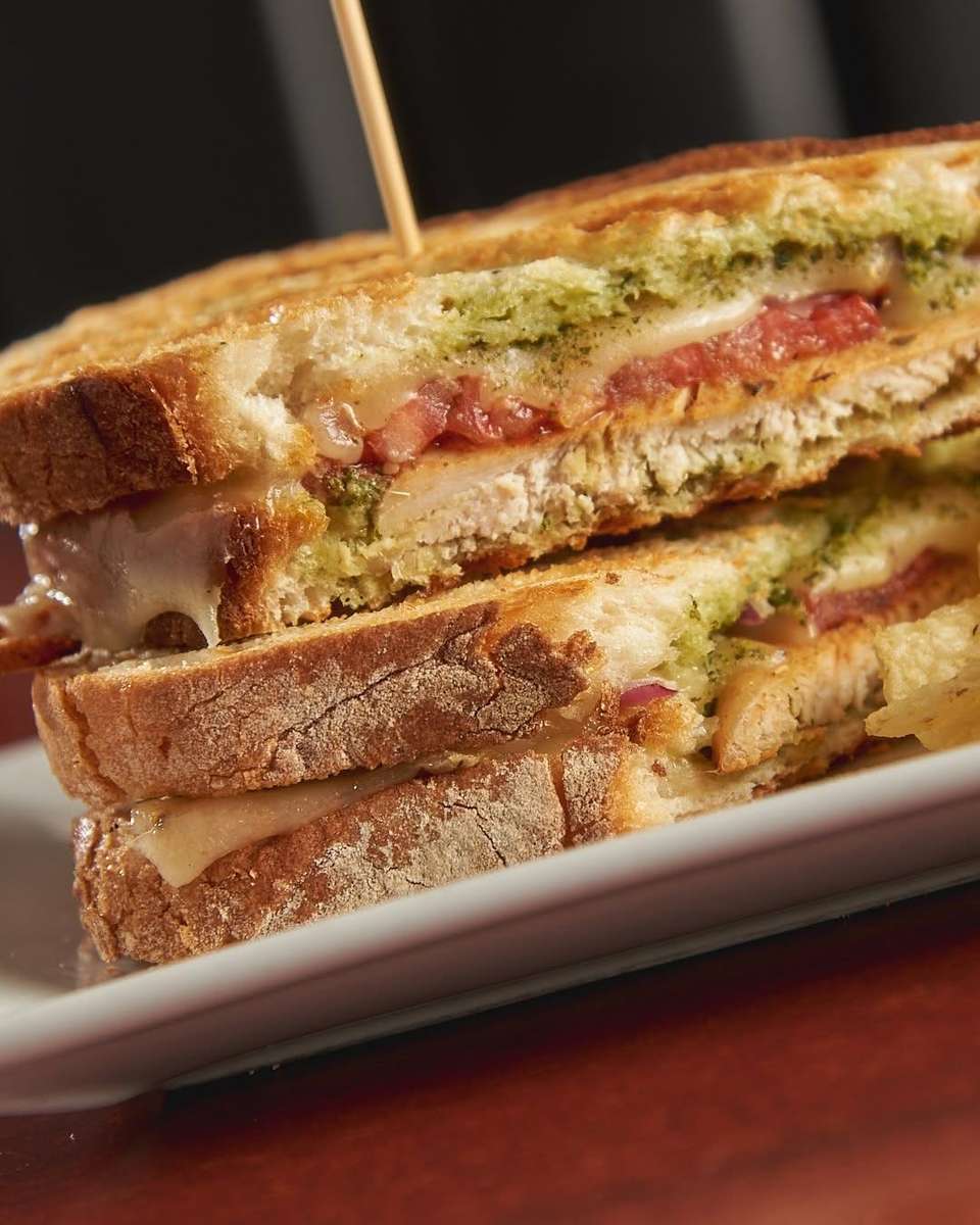 Close-up of a toasted sandwich layered with grilled chicken, melted cheese, tomato, pesto sauce, and red onion between slices of crusty artisan bread, served on a white plate for a hearty lunch option.