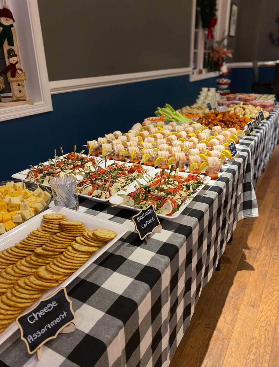 Elegant buffet table with a black-and-white checkered tablecloth featuring a festive appetizer spread including a cheese and cracker assortment, mozzarella caprese skewers, chilled shrimp cocktail with lemon wedges, buffalo wings, and vegetable crudités, set in a cozy indoor event space.