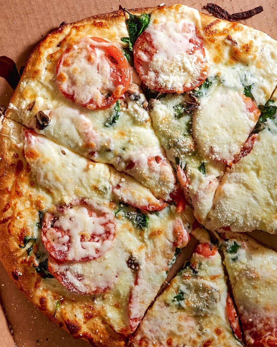 Close-up of a fresh Margherita pizza topped with melted mozzarella cheese, sliced tomatoes, fresh basil, and a sprinkle of grated Parmesan, served in a cardboard delivery box.