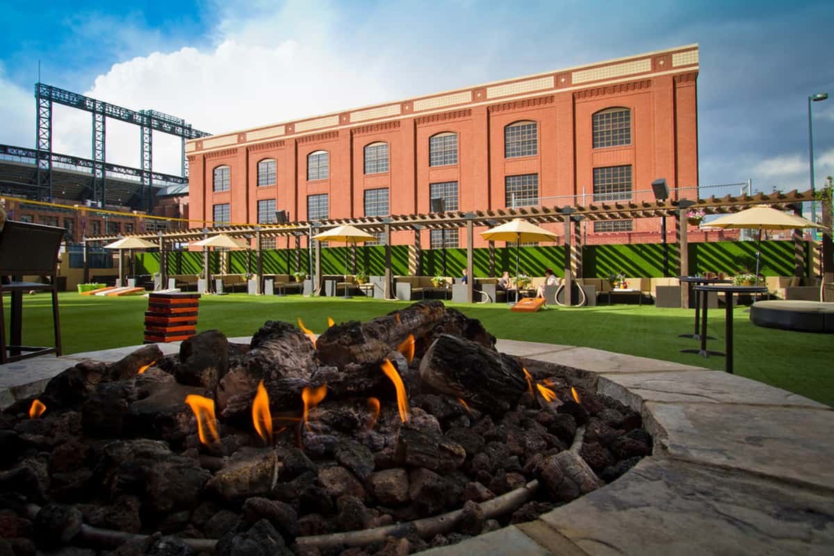Outdoor restaurant lawn with fire pit, pergola lounge seating, yard games, and open event space set against a downtown skyline backdrop.