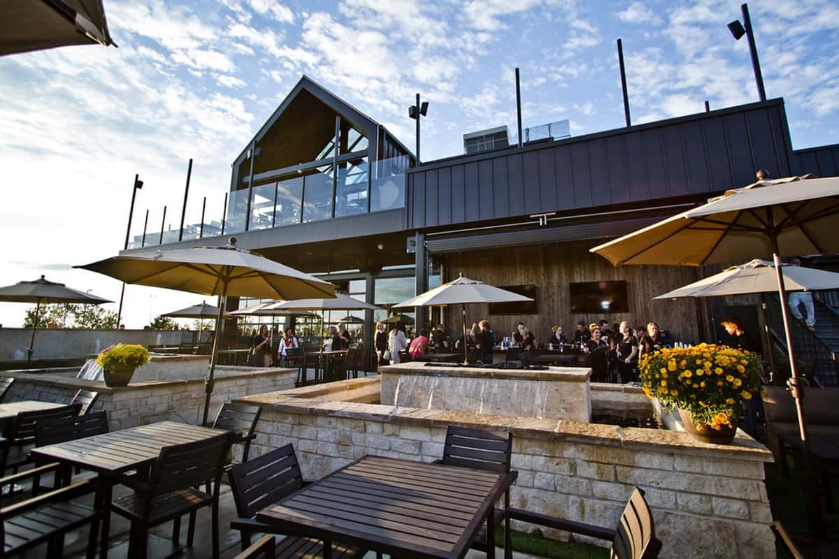 Modern restaurant patio with outdoor dining tables, umbrellas, fire features, and rooftop seating designed for social gatherings and scenic dining.