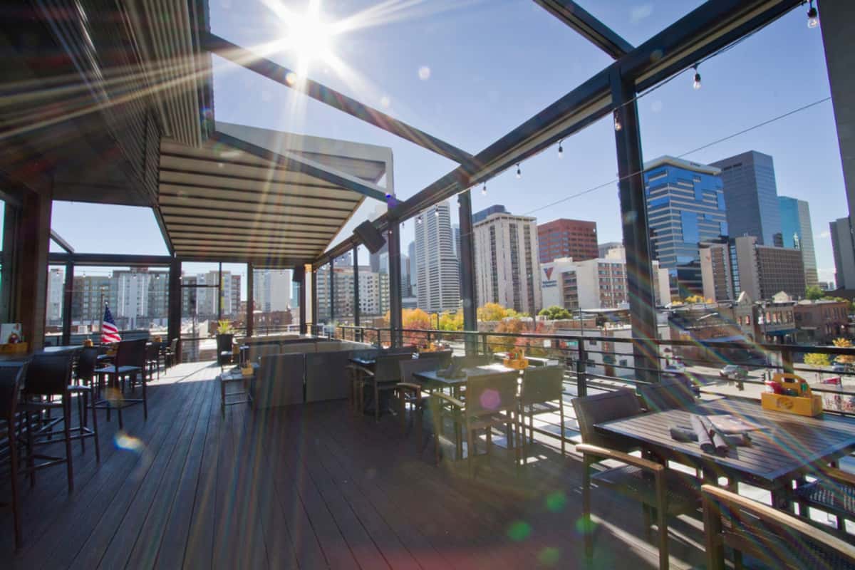 Sunlit rooftop patio with outdoor dining tables, modern lounge seating, and sweeping downtown skyline views.
