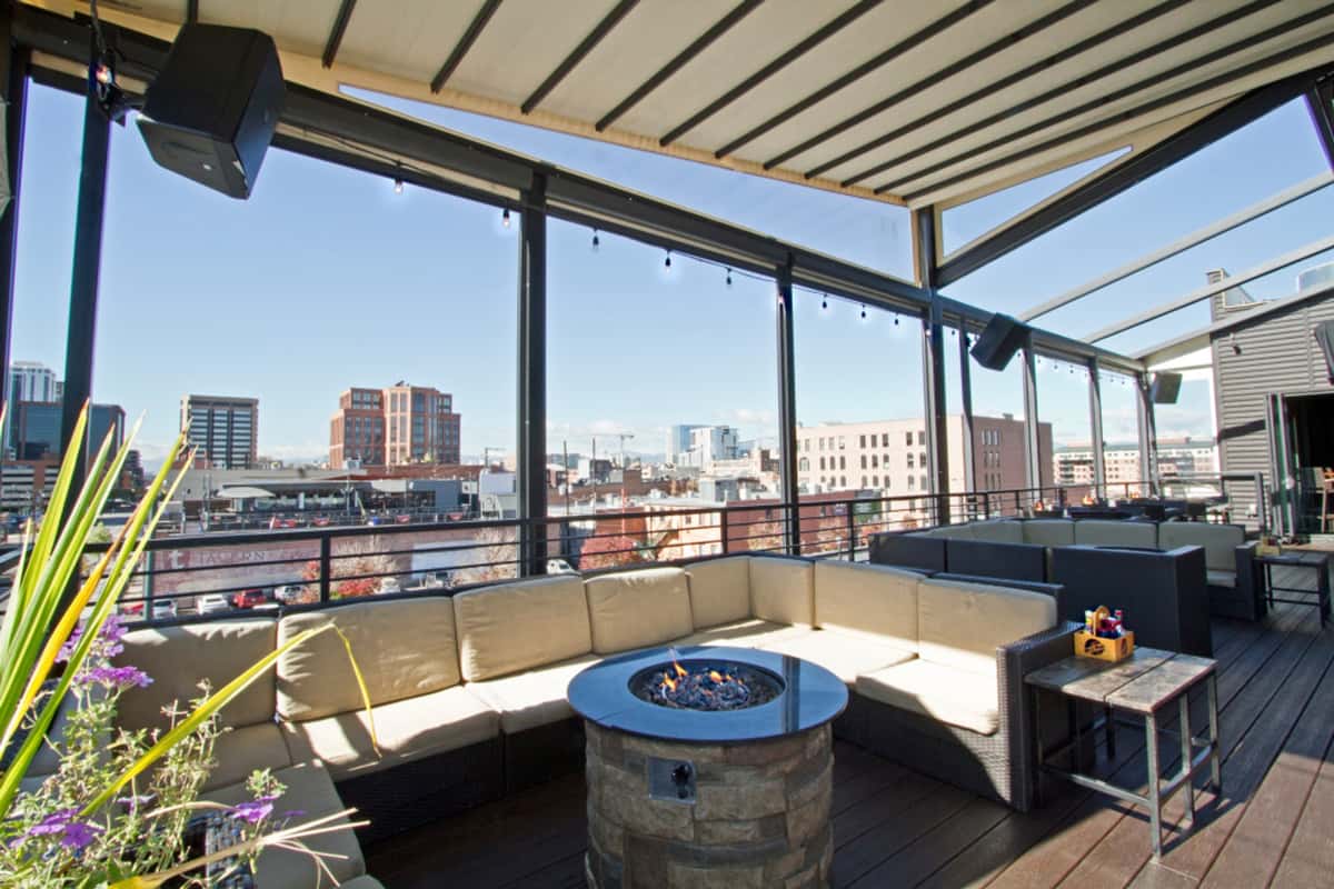 Rooftop patio lounge with sectional seating, fire pit table, and panoramic city skyline views in an upscale outdoor dining setting.