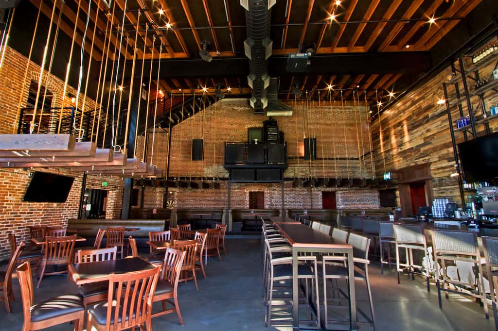 Large industrial-style restaurant and bar interior with exposed brick walls, communal seating, hanging light fixtures, and an open event-friendly layout.
