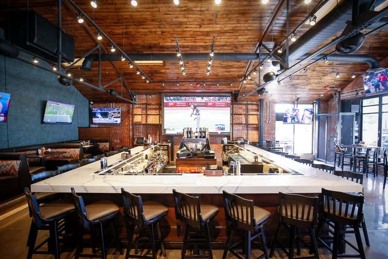 Industrial-style sports bar with a central island bar, multiple large-screen TVs, exposed brick walls, and communal seating for dining, drinks, and game-day gatherings.
