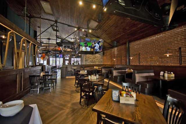 Warm industrial-style restaurant dining room with exposed brick walls, booth seating, wood tables, and televisions for casual dining and sports viewing.