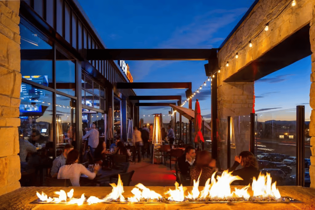 Outdoor rooftop patio at sunset featuring fire tables, string lighting, and skyline views for dining, cocktails, and social gatherings.