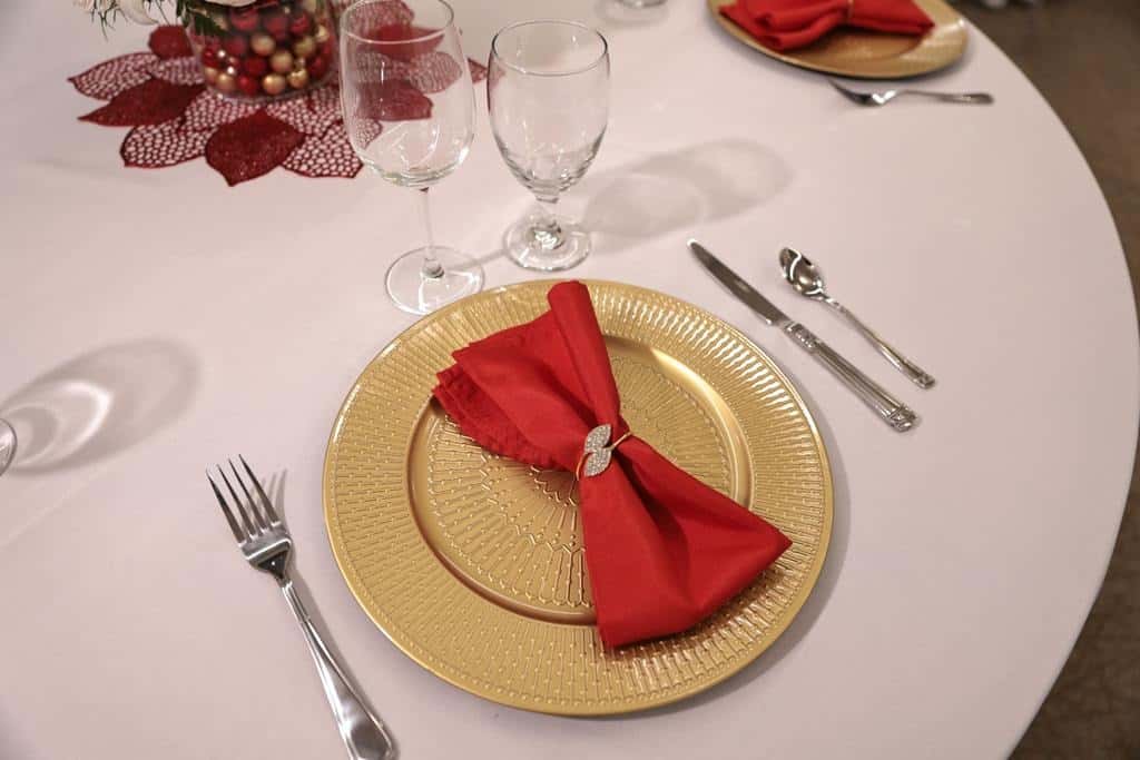 Elegant banquet table setting with a gold charger plate, neatly folded red napkin in a decorative ring, polished silverware, and wine glasses on a white tablecloth—festive fine-dining restaurant ambiance with upscale event décor and holiday-themed centerpiece.