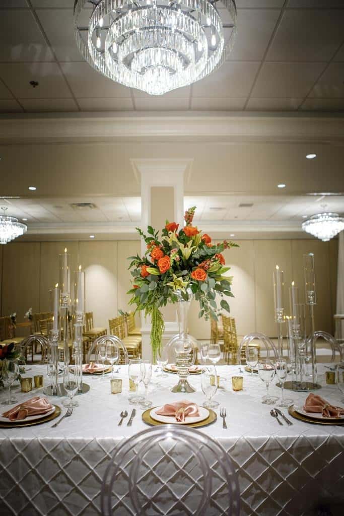 Elegant banquet table setting with crystal chandelier overhead, tall floral centerpiece with orange roses and greenery, candlelight, and gold-accented place settings in a luxury event or wedding reception venue