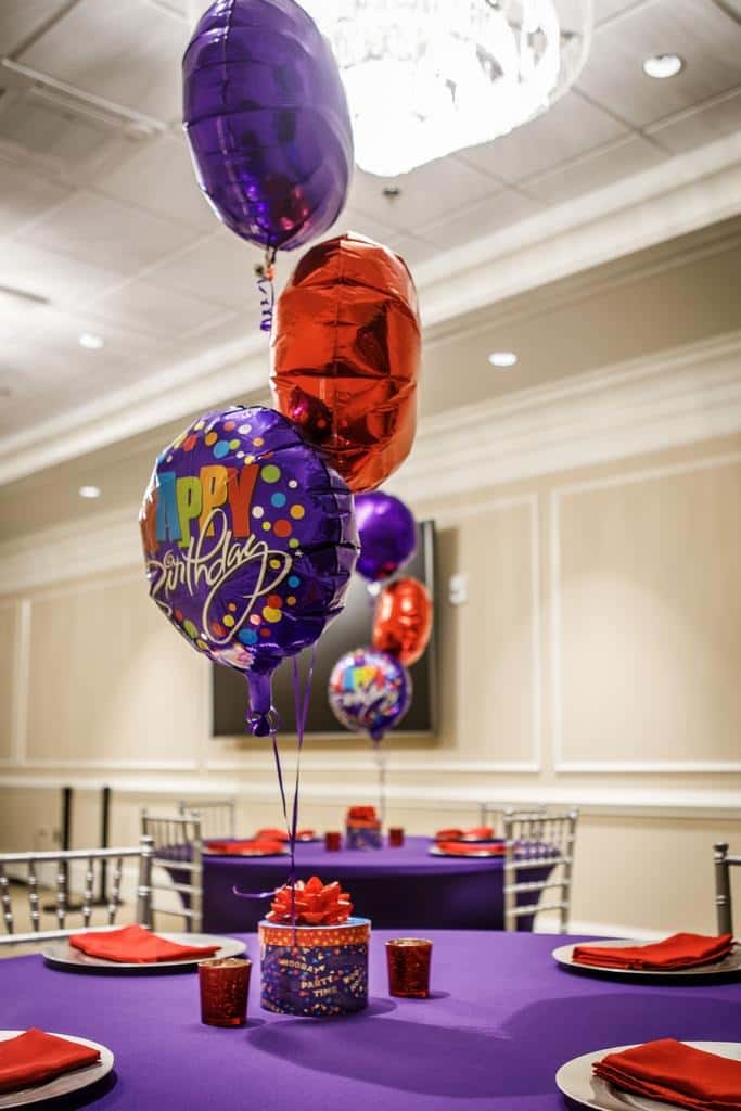 Birthday party event table setup with purple tablecloths, red napkins, and colorful helium balloons including a "Happy Birthday" balloon centerpiece—festive banquet hall décor for a catered birthday celebration or private party event.