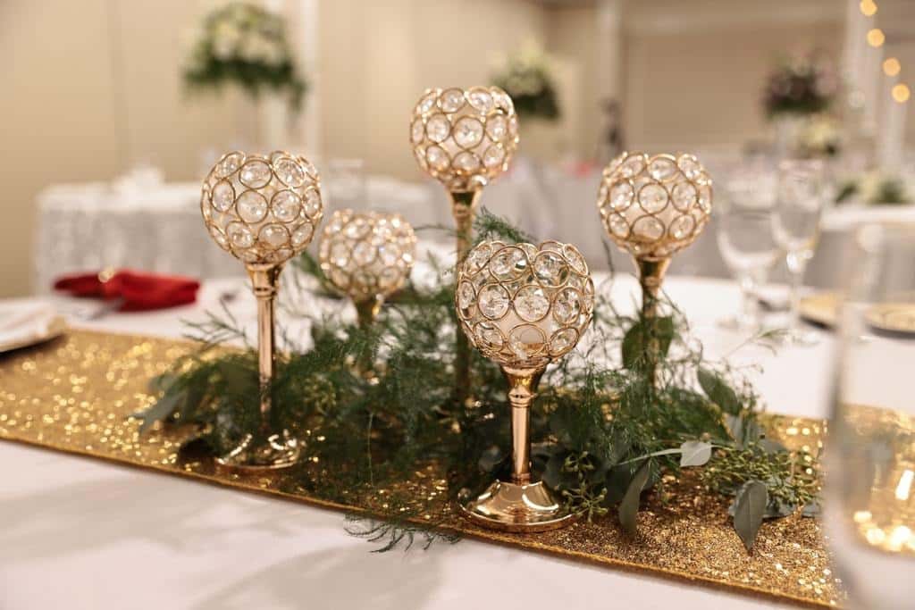Gold crystal candle holders with greenery centerpiece on a sparkling table runner, elegant wedding reception table décor in a luxury event venue.
