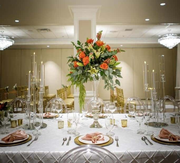 Elegant banquet table setting with crystal chandelier overhead, tall floral centerpiece with orange roses and greenery, candlelight, and gold-accented place settings in a luxury event or wedding reception venue