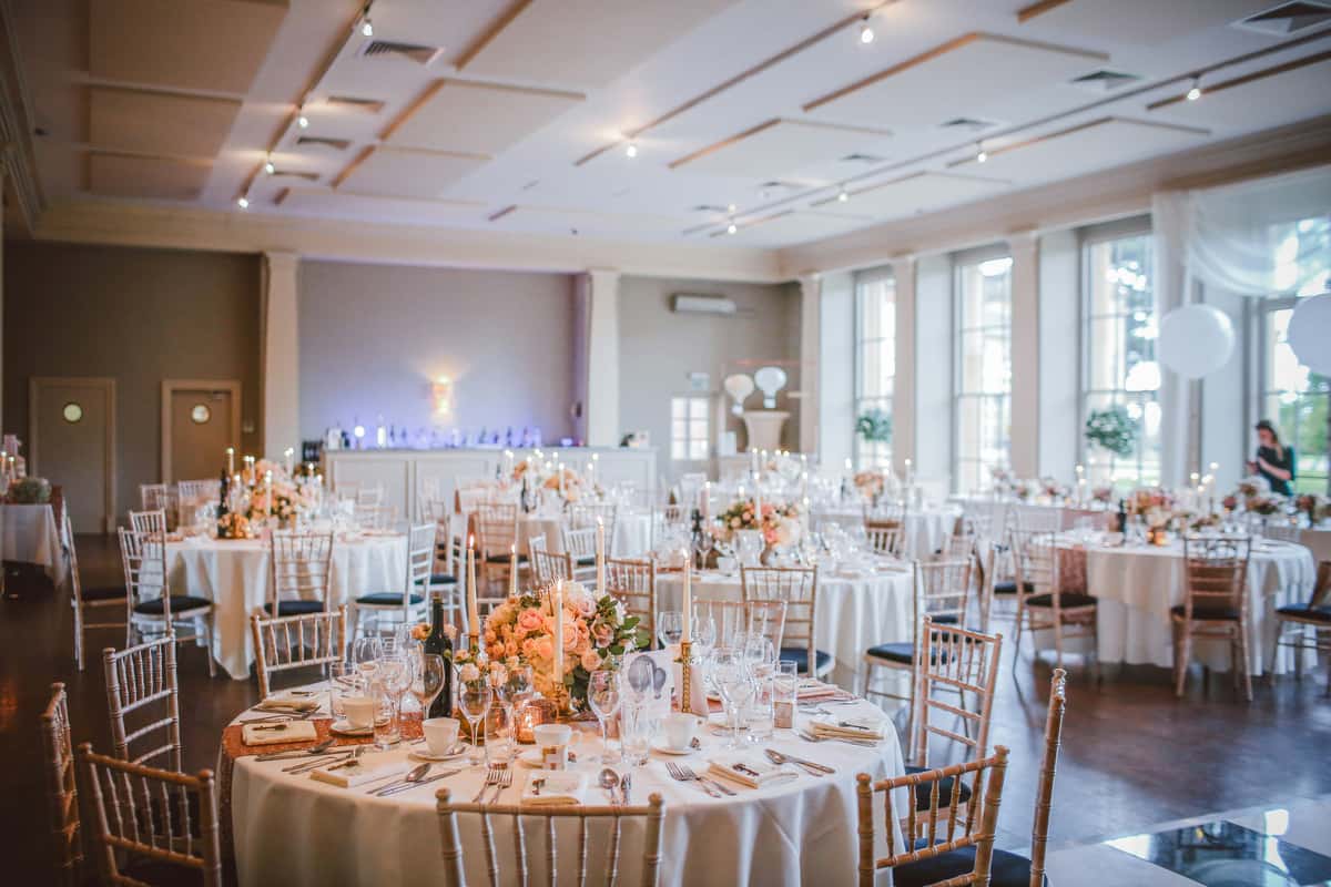Elegant wedding reception hall with round banquet tables, floral centerpieces, candles, and gold chairs set for a formal celebration.