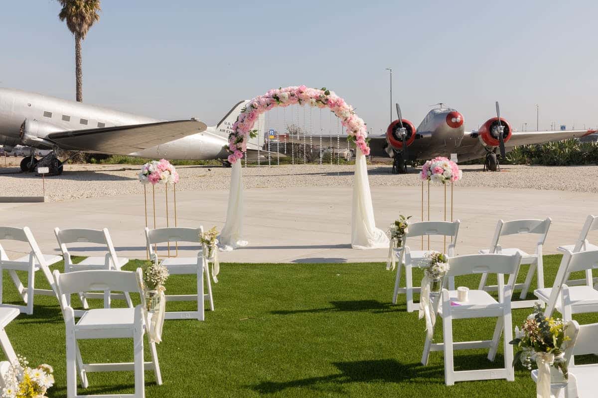 Outdoor wedding ceremony setup with floral arch, white folding chairs, and vintage airplane displays at an aviation-themed venue