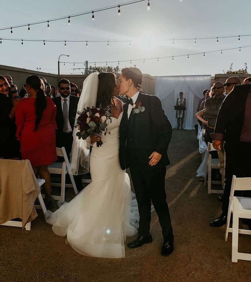 Bride and groom sharing a kiss during an outdoor wedding ceremony under string lights at a romantic sunset reception venue